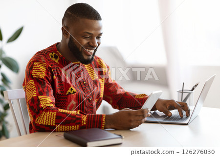 Cheerful Black Man In Traditional African Clothes Using Smartphone And Laptop While Working At Desk In Home Office, Smiling Millenial Guy Messaging On Cellphone, Enjoying Mobile Communication Cheerful Black Man In Traditional African Clothes Using Smartphone And Laptop While Working At Desk In Home Office, Smiling Millenial Guy Messaging On Cellphone, Enjoying Mobile Communication 126276030