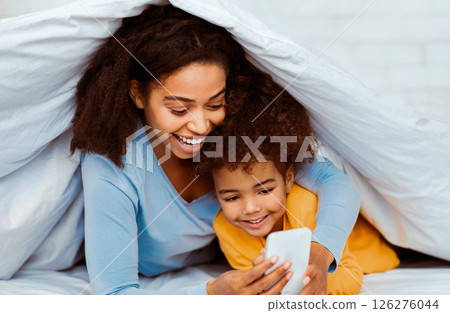 Happy Black Mother And Daughter Using Smartphone Lying In Bed Covered With Blanket In Bedroom. Selective Focus 126276044