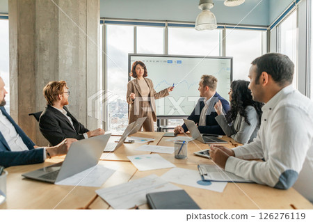 A businesswoman stands in front of a large screen and points to a graph as she presents data to a team of colleagues seated around a table in a modern office. A businesswoman stands in front of a large screen and points to a graph as she presents data to a team of colleagues seated around a table in a modern office. 126276119
