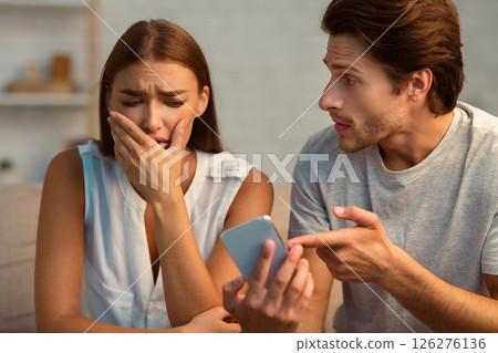 Cheating And Infidelity. Jealous Boyfriend Showing Phone To Crying Girlfriend Demanding Explanation Sitting On Couch Indoor. Selective Focus 126276136