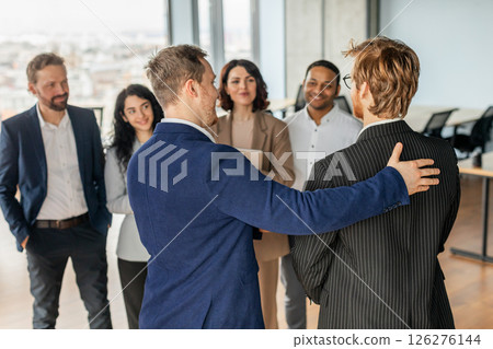 A group of professionals stand in an office setting, with one man in a blue suit placing a supportive hand on the shoulder of another man in a striped suit. 126276144