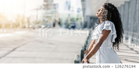 African American young woman with long, curly black hair stands on a city sidewalk, enjoying the sunshine and fresh air. She is wearing a white, striped dress with ruffled sleeves, copy space 126276194