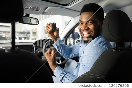 Own Car. Joyful African American Guy Holding Auto Key Shaking Fists Sitting In Automobile In Dealership Shop Own Car. Joyful African American Guy Holding Auto Key Shaking Fists Sitting In Automobile In Dealership Shop 126276206