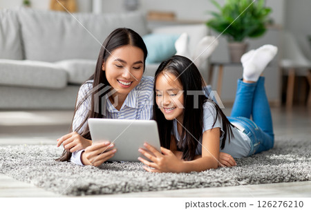 Excited asian mother helping her daughter with lesson on digital tablet, lying together on floor carpet in living room interior. Young mother and her child making online home assignment togeher Excited asian mother helping her daughter with lesson on digital tablet, lying together on floor carpet in living room interior. Young mother and her child making online home assignment togeher 126276210