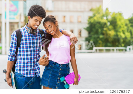 This image shows a teen black couple standing together on a city street. They are looking down at a smartphone and appear to be laughing, copy space 126276237