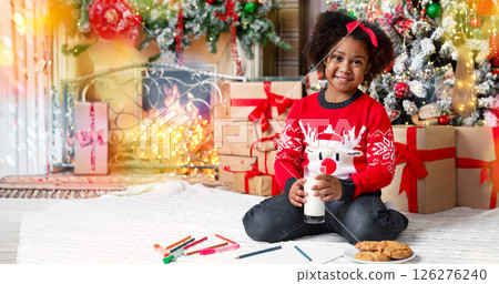 Adorable little black girl waiting for Santa with glass of milk and cookies, sitting near Christmas tree in decorated living room. 126276240