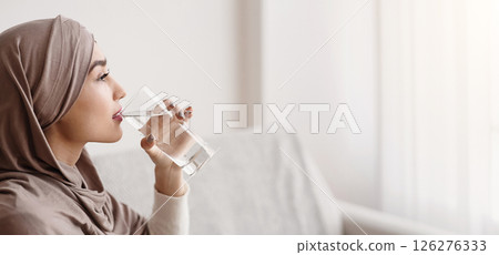 Thirsty islamic woman in headscarf drinking mineral water from glass at home, selective focus, empty space Thirsty islamic woman in headscarf drinking mineral water from glass at home, selective focus, empty space 126276333