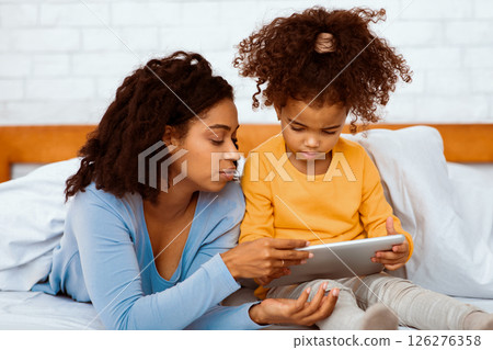 Black Mother Teaching Little Daughter To Use Digital Tablet Lying In Bed At Home. Computer Literacy For Kids 126276358