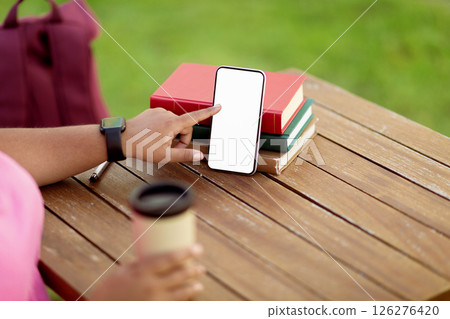 A person sits at a wooden table outdoors, pointing at a blank smartphone screen with one hand and holding a coffee cup with the other, surrounded by stacked books. A person sits at a wooden table outdoors, pointing at a blank smartphone screen with one hand and holding a coffee cup with the other, surrounded by stacked books. 126276420