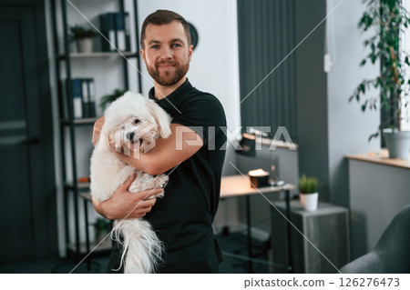 Modern office. Groomer is with Maltese dog indoors. Man is in the black uniform Modern office. Groomer is with Maltese dog indoors. Man is in the black uniform 126276473