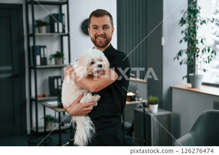 Modern office. Groomer is with Maltese dog indoors. Man is in the black uniform Modern office. Groomer is with Maltese dog indoors. Man is in the black uniform 126276474
