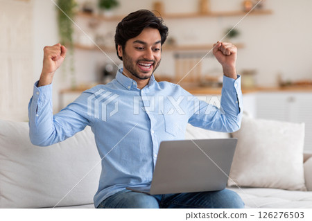 A young Arab man is sitting on a white sofa in a living room, looking at his laptop with a wide smile and raising both fists in the air in celebration. A young Arab man is sitting on a white sofa in a living room, looking at his laptop with a wide smile and raising both fists in the air in celebration. 126276503