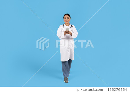 A healthcare professional Asian woman dressed in a white coat and scrubs walks confidently, engaged with a smartphone. The background is a vibrant blue, enhancing the professional's cheerful demeanor. 126276564