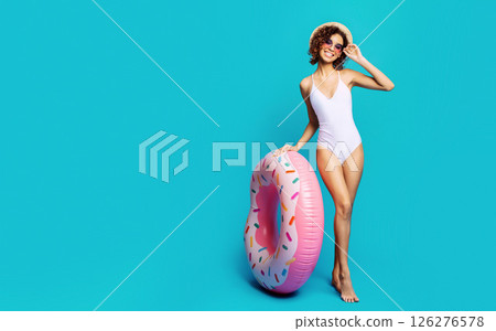 A woman in a white swimsuit and a straw hat stands with her left hand on a large pink inflatable donut float. She is smiling and wearing pink sunglasses. The background is a solid light blue color. A woman in a white swimsuit and a straw hat stands with her left hand on a large pink inflatable donut float. She is smiling and wearing pink sunglasses. The background is a solid light blue color. 126276578