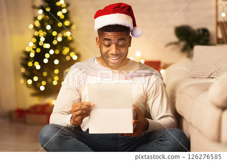 Christmas gift. Happy black guy opening box, sitting against Christmas tree and wearing santa hat 126276585