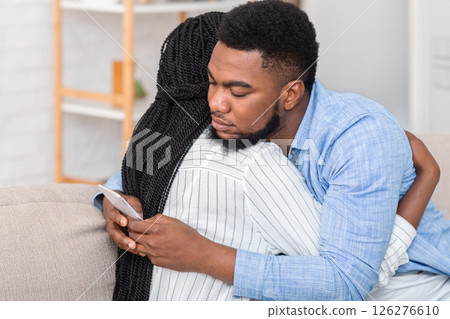 Cheating husband. Young african american man texting on cellphone with somebody while embracing his wife at home, closeup Cheating husband. Young african american man texting on cellphone with somebody while embracing his wife at home, closeup 126276610