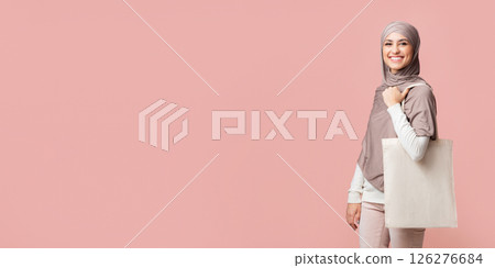Eco shopping. Cheerful arabic girl in hijab posing with tote canvas shopper bag with place for mockup, pink background Eco shopping. Cheerful arabic girl in hijab posing with tote canvas shopper bag with place for mockup, pink background 126276684