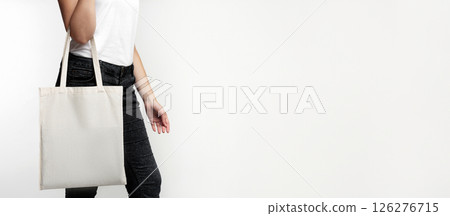 A woman in a white shirt and black jeans holds a plain white tote bag in front of a white background. 126276715
