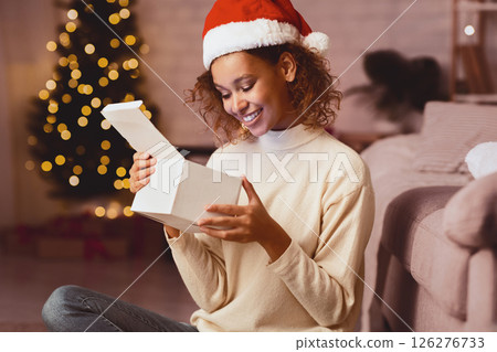 African-american girl looking inside Christmas present, sitting in front of xmas tree at home 126276733