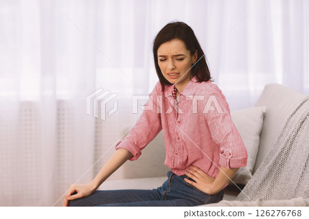 Side Stitch. Poor girl suffering from pain and cramps in her liver, sitting on sofa at home, holding spine, copyspace Side Stitch. Poor girl suffering from pain and cramps in her liver, sitting on sofa at home, holding spine, copyspace 126276768