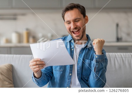 A young man sits on a couch in his home, reading a piece of paper with a big smile on his face. He is excited and holding his fist in the air. He is wearing a blue shirt over a white t-shirt. A young man sits on a couch in his home, reading a piece of paper with a big smile on his face. He is excited and holding his fist in the air. He is wearing a blue shirt over a white t-shirt. 126276805