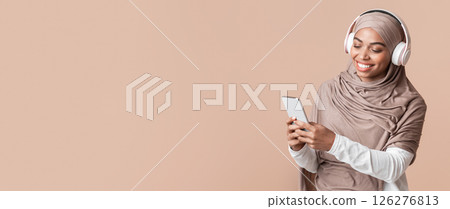 Cool music app. Smiling black muslim girl in hijab listening her favorite songs with wireless headphones online on smartphone Cool music app. Smiling black muslim girl in hijab listening her favorite songs with wireless headphones online on smartphone 126276813
