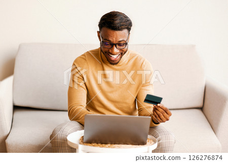 Online Shopping. Excited Black Guy Using Credit Card And Laptop Computer Sitting On Sofa Indoor. Selective Focus Online Shopping. Excited Black Guy Using Credit Card And Laptop Computer Sitting On Sofa Indoor. Selective Focus 126276874