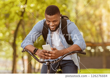 Outdoor portrait of smiling young afro man with mobile phone in the street, sitting on bike, downloading music playlist, riding to work 126276884