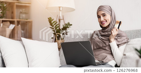 Shopping online is easy. Portrait of happy muslim woman in hijab with laptop and credit card sitting on sofa at home and smiling at camera Shopping online is easy. Portrait of happy muslim woman in hijab with laptop and credit card sitting on sofa at home and smiling at camera 126276968