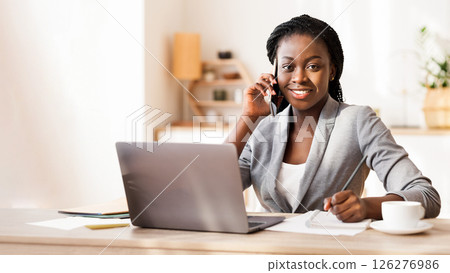 Portrait of black female entrepreneur talking on mobile phone, taking notes and smiling to camera 126276986