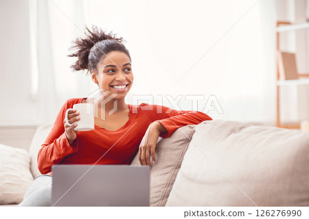 Smiling African American Lady With Laptop Having Coffee Sitting On Sofa At Home. Enjoying Weekend Morning. Empty Space Smiling African American Lady With Laptop Having Coffee Sitting On Sofa At Home. Enjoying Weekend Morning. Empty Space 126276990