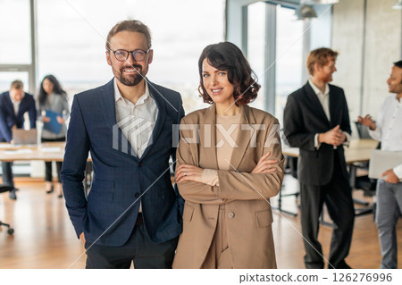 A man and woman in business attire stand confidently in front of a large window. They are both smiling and appear to be happy and successful. The background is blurred and features other people 126276996