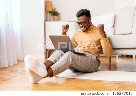 Excited African American Guy Holding Laptop Shaking Fists Celebrating Success Sitting On Floor Indoor. Selective Focus Excited African American Guy Holding Laptop Shaking Fists Celebrating Success Sitting On Floor Indoor. Selective Focus 126277015