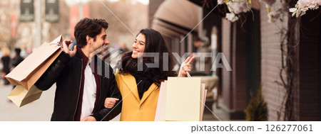 A young couple walks together, laughing and carrying shopping bags on a vibrant city street adorned with spring flowers, copy space A young couple walks together, laughing and carrying shopping bags on a vibrant city street adorned with spring flowers, copy space 126277061