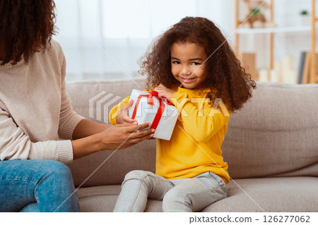 Kid's Birthday. Cute African Little Girl Unpacking Wrapped Gift Sitting With Mother On Sofa At Home. Kid's Birthday. Cute African Little Girl Unpacking Wrapped Gift Sitting With Mother On Sofa At Home. 126277062