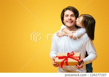 Cheerful little daughter kissing her birthday father, dad holding gift box, Father Day concept, yellow background 126277107