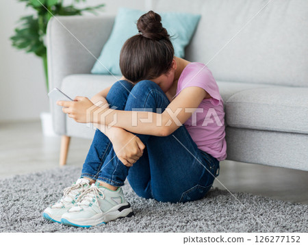 Depressed Indian teen girl with cellphone crying on floor at home, suffering from cyber bullying, being harassed online. Stressed teenager being victim of internet abuse or trolling 126277152