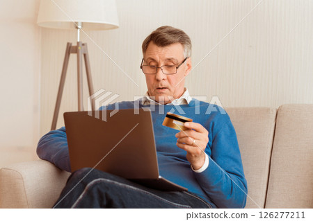 Online Shopping Concept. Elderly Gentleman Using Credit Card And Laptop Computer Buying Things On Internet Sitting On Sofa At Home Online Shopping Concept. Elderly Gentleman Using Credit Card And Laptop Computer Buying Things On Internet Sitting On Sofa At Home 126277211