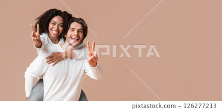 Joyful interracial couple embracing and showing peace sign, posing together over yellow background 126277213