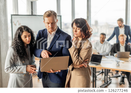 A group of business professionals are gathered around a table in a modern office setting. One man holds a laptop open, pointing at the screen while talking to women standing beside him. A group of business professionals are gathered around a table in a modern office setting. One man holds a laptop open, pointing at the screen while talking to women standing beside him. 126277246