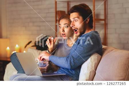Shocked Young Couple Using Laptop Watching Movie Eating Popcorn Sitting On Couch At Home At Night. Selective Focus Shocked Young Couple Using Laptop Watching Movie Eating Popcorn Sitting On Couch At Home At Night. Selective Focus 126277265