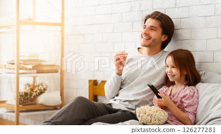 Lazy weekend at home. Cute little girl watching TV with her young daddy, eating popcorn, copy space Lazy weekend at home. Cute little girl watching TV with her young daddy, eating popcorn, copy space 126277281