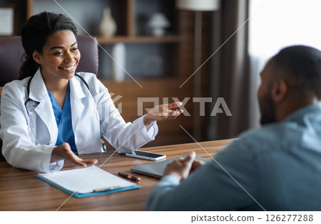 Friendly attractive african american young woman doctor having conversation with male patient, general practitioner consulting young man, gesturing and smiling, giving details, modern clinic interior Friendly attractive african american young woman doctor having conversation with male patient, general practitioner consulting young man, gesturing and smiling, giving details, modern clinic interior 126277288