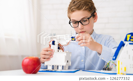 A young boy sits at a table, wearing glasses, and is deeply focused on assembling a robotics project. He holds a small component in his hand, carefully aligning it with the rest of the construction A young boy sits at a table, wearing glasses, and is deeply focused on assembling a robotics project. He holds a small component in his hand, carefully aligning it with the rest of the construction 126277332