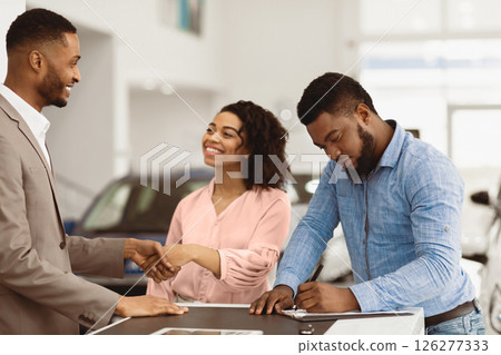 Buying New Car. African American Spouses Shaking Hands With Auto Seller Signing Papers In Dealership Office. Selective Focus Buying New Car. African American Spouses Shaking Hands With Auto Seller Signing Papers In Dealership Office. Selective Focus 126277333