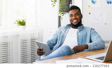 Ready for presentation. Portrait of black millennial employee holding digital tablet, sitting at workplace and looking at camera Ready for presentation. Portrait of black millennial employee holding digital tablet, sitting at workplace and looking at camera 126277363