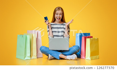 Big sales. Excited girl holding laptop and credit card, sitting on floor surrounded by bright paper bags on orange studio background 126277370