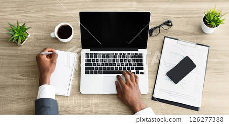 A person sits at a desk, working on a laptop while taking notes on a notepad. 126277388