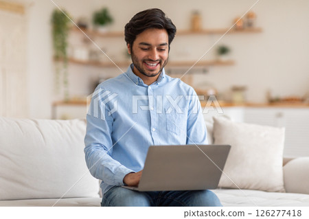 Arab man with short dark hair, wearing a light blue button-down shirt and jeans, sits on a white couch in a living room and uses a silver laptop. He is looking at the screen and smiling Arab man with short dark hair, wearing a light blue button-down shirt and jeans, sits on a white couch in a living room and uses a silver laptop. He is looking at the screen and smiling 126277418