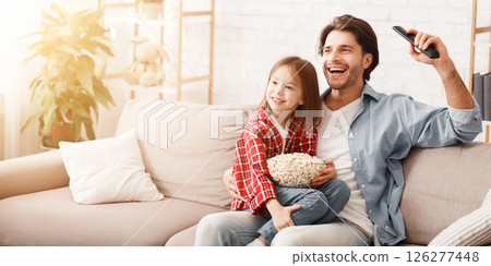 Little girl and her happy father watching football match on TV, celebrating success of their team, sitting on sofa with popcorn, family traditions concept, copy space 126277448